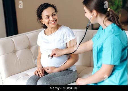 Donna incinta felice e medico femminile. Medico amichevole esamina gravidanza mista razza donna sorridente, ascoltando il battito cardiaco del bambino, seduta su un divano a casa o ospedale, sano concetto di gravidanza Foto Stock