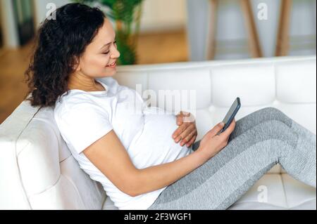 Donna soddisfatta in attesa di un bambino. Razza mista buona donna incinta adulta passare il tempo libero utilizzando smartphone, navigare in internet, chattare online con amici o familiari, si siede sul divano a casa Foto Stock