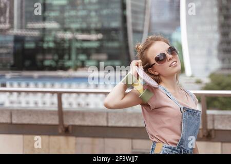 Stile di vita estivo di bella ragazza alla moda con penny board rosa su sfondo grattacieli. Indossare occhiali da sole eleganti e tute in denim Foto Stock