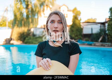 Ritratto di una donna elegante guarda in su, stellata, tenendo il suo cappello nelle sue mani. Sullo sfondo c'è una piscina e una casa. Concetto di emozione umana Foto Stock