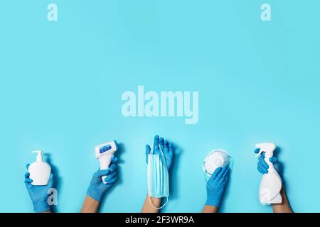 Mani sollevate in guanti medici che tengono maschere, disinfettanti, sapone, termometro senza contatto su sfondo blu. Banner. Spazio di copia. Protezione della salute Foto Stock