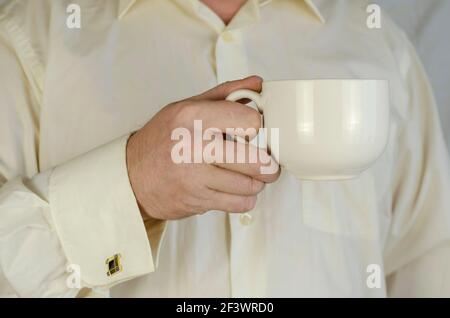 L'uomo adulto sta tenendo una grande tazza bianca. Un uomo caucasico di mezza età in una camicia crema con gemelli. All'interno. Messa a fuoco selettiva. Foto Stock