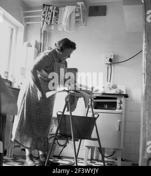 1950s, storica, una donna matura in una pinafore in una cucina utilizzando un semplice manle per estrarre l'acqua dai vestiti lavati, Inghilterra, Regno Unito, con un secchio di metallo sotto per catturare l'acqua in eccesso. Un maniglione o strizzatoio, come è anche noto, è un sussidio meccanico per biancheria, che presenta due rulli in un telaio ed è lavorato da una manovella a mano per strizzarne l'acqua dalla biancheria umida. Foto Stock