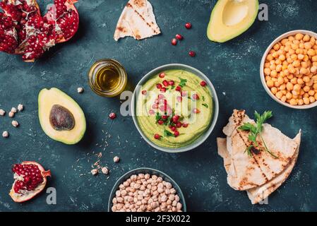 Cucina mediorientale, ebraica, arabica. Hummus verde fatto in casa con avocado, prezzemolo, olio d'oliva, pita, ceci crudi, melograno su sfondo scuro Foto Stock