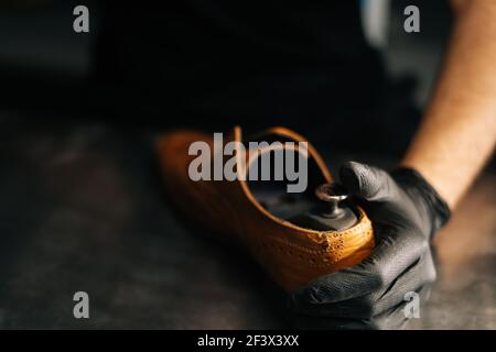 Mani ravvicinato di calzolaio indossando guanti neri inserti in legno le scarpe si bloccano nelle scarpe in pelle marrone chiaro indossate Foto Stock