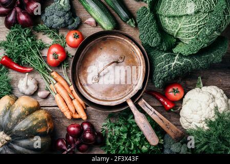 Pentola di rame, erbe, cipolla viola, zucca, pomodoro, broccoli, pepe, cavolo, carota, prezzemolo, calafiore, zucchine su fondo ligneo. Vista dall'alto. Copia Foto Stock