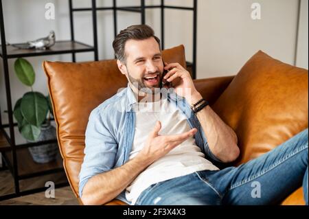 Ragazzo giovane e rilassato, sopportato da millenni, sdraiato sul divano nel salotto, tenendo il telefono cellulare, gesturando mentre si parla con l'amico, discutendo di progetto, pettegolare, fare scherzi, comunicare Foto Stock