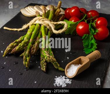 Composizione di verdure varie con mazzo di asparagi, funghi, pomodori ciliegini e prezzemolo, con cucchiaio di legno contenente sale sulla superficie dell'ardesia Foto Stock