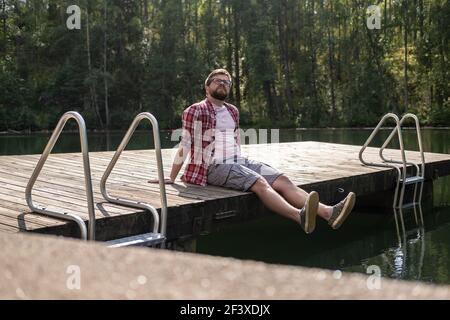 L'uomo calmo siede sul bordo di un molo di legno e guarda in su pensieroso, sullo sfondo di un lago di foresta calmo. Stile di vita. Foto Stock