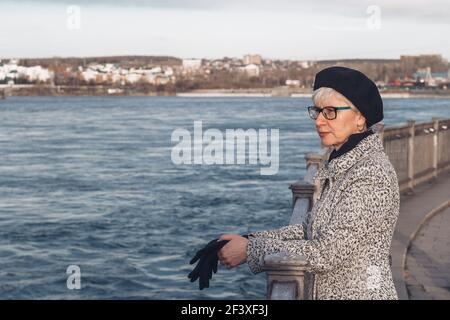 Ritratto di un'elegante donna di mezza età in occhiali sull'argine del fiume. Uno sguardo penoso nella distanza.solitudine, riflessione, rilassamento Foto Stock