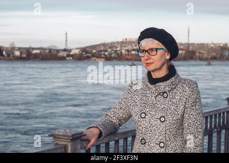 Ritratto di un'elegante donna di mezza età in occhiali sull'argine del fiume. Primo piano. Uno sguardo penoso sulla distanza. Solitudine, riflessione, Foto Stock