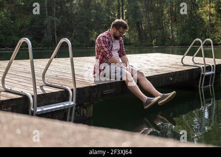 L'uomo sconvolto si siede sul bordo di un molo di legno e gazes pensieroso nell'acqua. Stile di vita. Foto Stock