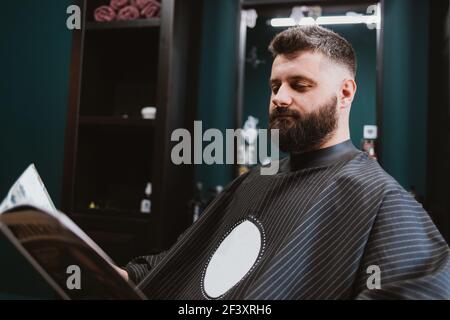 Barbiere cliente, giovane con barba, seduta in poltrona e rivista di lettura. Foto Stock