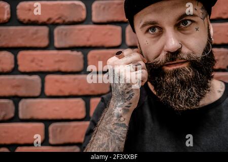 Ritratto di un bel hipster vecchio stile in t-shirt e cappuccio, che tocca la barba. Su uno sfondo di mattone. Foto Stock