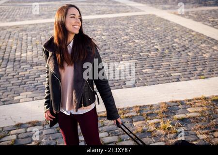 giovane donna sorridente con valigia trolley in una strada acciottolata, concetto di viaggio e stile di vita urbano, copyspace per il testo Foto Stock