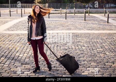 felice giovane donna con trolley valigia a piedi lungo una strada acciottolata, concetto di viaggio e stile di vita urbano, copyspace per il testo Foto Stock