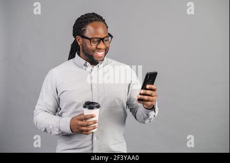 Uomo d'affari afro-americano impegnato e sicuro che indossa una camicia casual elegante e occhiali che testano sullo smartphone e che tiene una tazza di caffè da asporto, dipendente dell'ufficio maschile isolato in grigio Foto Stock