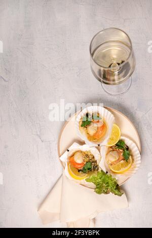 Capesante al forno con caviale in un piatto e un bicchiere di vino bianco. Capesante con limone su fondo nero con tovagliolo e forchetta. Foto Stock