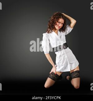 giovane bella donna in abito corto bianco in posa in studio inginocchiarsi su sfondo scuro Foto Stock