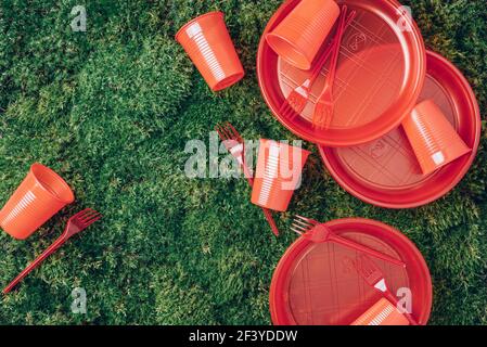 Inquinamento ambientale. Stoviglie in plastica monouso - piastre in plastica, forcelle, cucchiai, coltelli. Stoviglie di plastica rossa su erba verde, fondo muschio Foto Stock