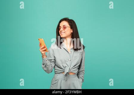Foto di bella signora in occhiali da sole tiene smartphone. Indossa una camicia grigia, uno sfondo isolato di colore turchese Foto Stock