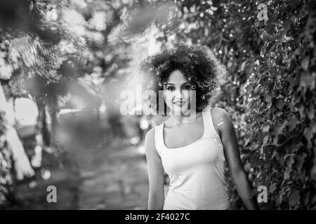 Giovane donna nera con acconciatura afro sorridente nel parco urbano. Giovane donna nera con acconciatura afro sorridente nel parco urbano. Ragazza mista che indossa abiti casual. Fotografia in bianco e nero. Foto Stock