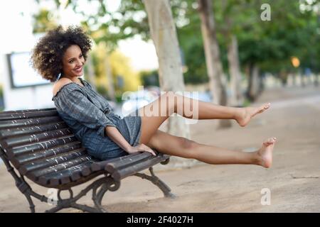 Giovane donna nera con acconciatura afro sorridente in backgroun urbano. Giovane donna nera con acconciatura afro seduta su una panca in fondo urbano che le muove le gambe. Ragazza mista che indossa abiti casual. Foto Stock