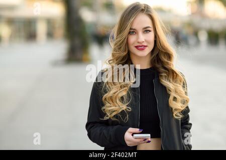 Donna bionda texting con il suo smart phone in background urbano. Bellissima fanciulla indossando giacca nera a piedi in strada. Pretty donne russe con lunghi capelli mossi di acconciatura. Foto Stock