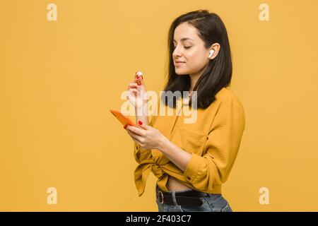 Primo piano foto di giovane donna sta utilizzando EarPods e smartphone. Indossa una camicia gialla, isolato di colore giallo Foto Stock