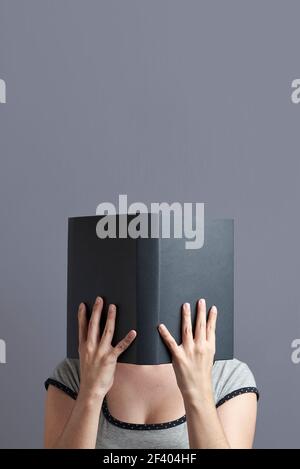 Giovane donna con il suo volto nascosto dietro un libro nero che tiene con entrambe le mani, aperto, la lettura. Verticale con sfondo grigio e spazio per la copia. C Foto Stock