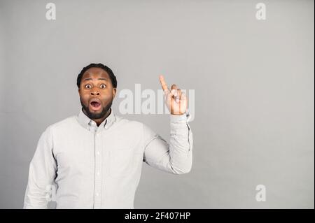Eccitato afroamericano ragazzo è venuto con una nuova soluzione, un biracial sorpreso uomo che indossa smart camicia casual punta dito in su, spazio vuoto di copia per la pubblicità di testo, isolato su grigio Foto Stock