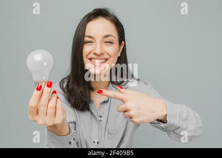 Foto di simpatica femmina sorridente tiene la lampada e punta verso di essa. Indossa un abito grigio, isolato sfondo di colore grigio Foto Stock