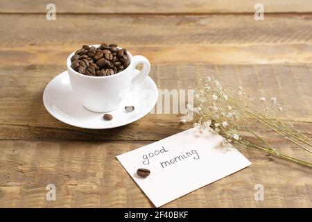 Chicchi di caffè in una tazza bianca con un cartoncino che riporta la scritta "Buongiorno!" su sfondo di legno. Concetto di caffè del mattino. Foto Stock
