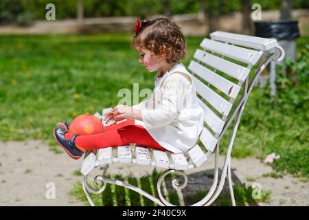 Adorabile bambina gioca con una sfera seduta su una panchina nel parco Foto Stock