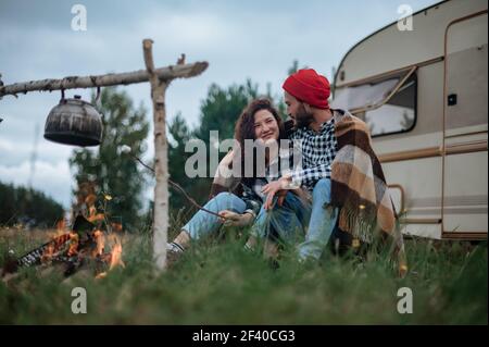 Accoppia in un plaid a scacchi arrostendo marshmallows sul fuoco vicino alla casa del rimorchio. Foto Stock