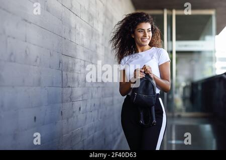 Giovane donna africana con nero capelli ricci passeggiate vicino edificio aziendale. Felice ragazza araba in sport vestiti in strada. Foto Stock