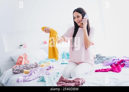 Foto di carino giovane donna nightwear seduto letto disordine vestiti scegliendo che cosa mettere sul dispositivo moderno all'interno camera in casa Foto Stock