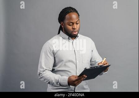 Concentrato afroamericano giovane uomo con dreadlock tenendo una cartella, prendendo appunti, messo a fuoco uomo nero scrive con penna, isolato su sfondo grigio Foto Stock