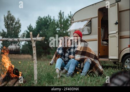 Accoppia in un plaid a scacchi arrostendo marshmallows sul fuoco vicino alla casa del rimorchio. Foto Stock