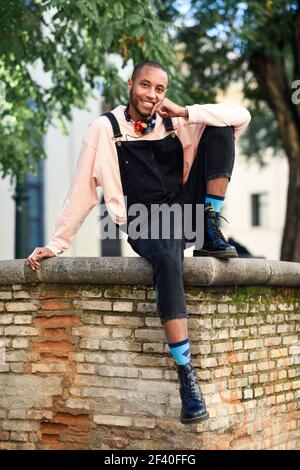 Giovane uomo nero di indossare un abbigliamento informale e cuffie seduti sul background urbano. Felice ragazzo africano con bib pants all'aperto Foto Stock