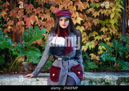 Giovane bella ragazza con peli molto lunghi che indossa cappotto invernale e cappuccio in foglie di autunno sfondo. Lo stile di vita e il concetto di moda. Foto Stock