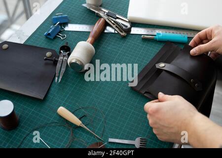 Mano uomo che lavora sul portafoglio in pelle nella sua officina. Lavorazione con pelle naturale marrone. Artigiano che tiene un attrezzi di lavorazione. Foto Stock