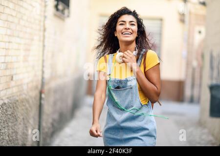 Giovane donna africana con cuffie e capelli neri ricci che camminano all'aperto. Ragazza felice che indossa t-shirt gialla e abito in denim su sfondo urbano. Giovane donna africana con cuffie che cammina all'aperto Foto Stock