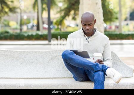 Giovane uomo nero con computer tablet e take away caffè in ambiente urbano. Ragazzo africano giovane con la testa rasata che indossa abiti casual. Giovane uomo nero con computer tablet e caffè da portare via Foto Stock