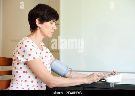 Donna di mezza età che misura la propria pressione sanguigna con un dispositivo elettronico per la misurazione della pressione sanguigna a casa. Donna che misura la propria pressione sanguigna a casa. Foto Stock