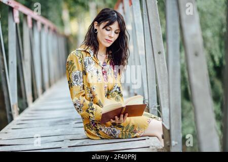 Giovane donna, vestita con fiori, che legge un libro su un ponte rurale. Giovane donna che legge un libro su un ponte rurale. Foto Stock