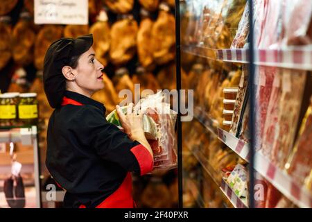 Ritratto di operaio femminile che prende i prodotti nel macellaio. Vetrina refrigerata per confezioni di salsicce, prosciutto e formaggio. Ritratto di operaio femminile che prende i prodotti nel macellaio Foto Stock