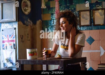 Giovane donna arabica con gesto premuroso seduta in un bel bar con decorazione vintage. Ragazza araba in abiti casual che beve una soda.. Donna araba in un bel bar che guarda attraverso la finestra Foto Stock