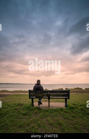 Vista posteriore di una donna seduta su una panchina, che guarda verso il mare, cielo di moody. Foto Stock