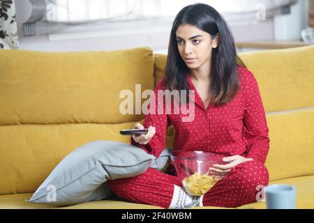 Donna persiana a casa che guarda la TV e usa il telecomando. Ragazza mangiare patatine patate. Donna persiana a casa che guarda la TV e usa il telecomando Foto Stock